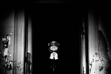 Dramatic black and white photo in the style of steam punk of a blonde girl in steampunk glasses and a black hoodie with a hood in the doorway of an abandoned old building