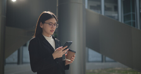 Fototapeta premium Young Asian business woman in suit holding mobile phone with takeaway coffee