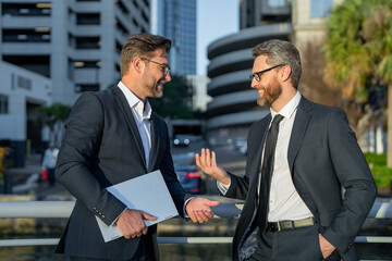 Business communication. Two business man in suit talking about finance news standing in modern city. Businessmen walking and talking outdoor in New York city. Confident employee talk bysiness idea.