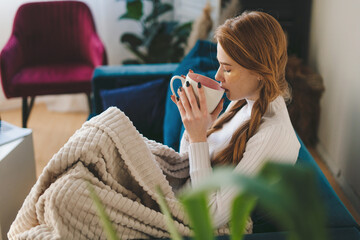 Dreamy satisfied woman wrapped warm blanket relaxing at home, sitting on cozy couch, drinking tea...