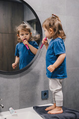 Caucasian little girl brushing teeth in bathroom looking in mirror while using toothbrush with...