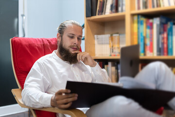 ฺBusinessman caucasian with beard using laptop. Sitting and work in book corner. Male manager raised hand to touch head thinking planning business ideas in library with bookshelves. Relax time.