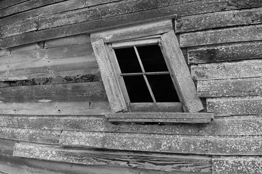 Black and white photo of window in weathered, old, abandoned barn. Window pane broken and missing, frame tilted.