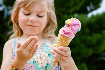 Happy preschool girl eating colorful ice cream in waffle cone on sunny summer day. Little toddler child eat icecream dessert. Sweet food on hot warm summertime days. Bright light, colorful ice-cream