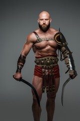 Bald and bearded gladiator poses standing tall with two swords while wearing light armor. This fearsome and imposing warrior emanates power and strength against a neutral gray background