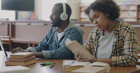 Two African American students sitting at desk in library listening to music through headphones and reading book. Campus lifestyle and people concept.