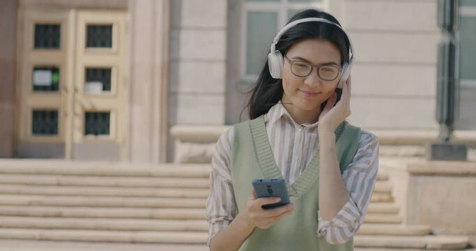 Slow motion portrait of happy Asian woman listening to music with headphones holding smartphone walking in city on sunny summer day