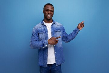 handsome young american man in denim jacket showing hand to side on blue background with copy space