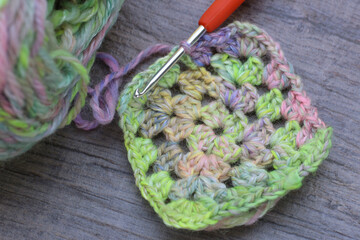 Closeup of craft work of crochet granny squares in mute rainbow colours with a crochet hook....