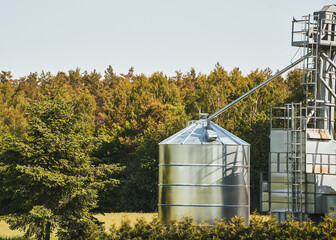 Steel grain silos stand next to a field. Agro-Industrial landscape. Steel Silos Embrace Vast Fields, Serving as Epitomes of Agricultural Storage and Processing © AlexGo