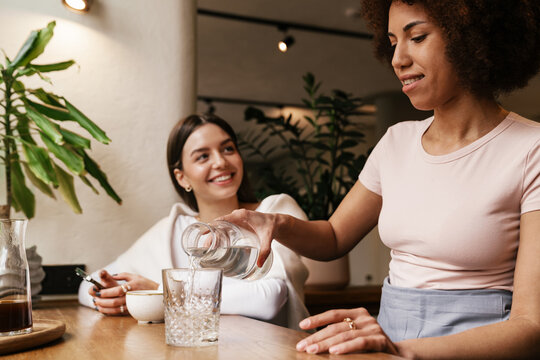 Smiling waitress pouring water in glass while serving for woman in cafe