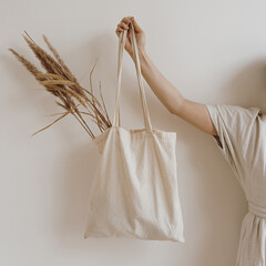 Young beautiful woman in neutral beige creamy linen dress holding sandy shopper bag with dried pampas grass over white wall. Mock up with blank copy space. Shopping