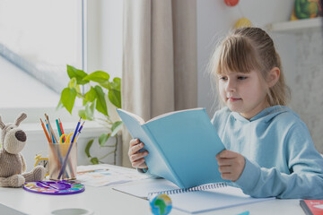 Schooler cute little girl sitting at desk in bedroom, holding a book and reading literature, doing homework, kid studying at home, getting ready before exams. Home education, homeschooling