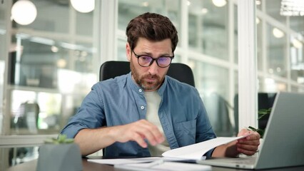 Tired exhausted businessman office worker in glasses sits at his desk reading documents. Male specialist is tired of the routine work indoors and wants to take a break, feeling a headache.