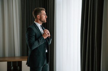 Fixes the tie. Businessman is indoors in the hotel room