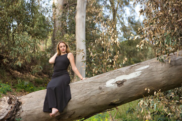 Young woman, blonde and beautiful, with a black dress, sitting on the trunk of a large tree, in the middle of nature, pure and virginal. Concept nature, peace, purity, virginity, trees.