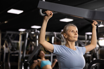 Fototapeta na wymiar Focused aged woman working out back muscles in modern gym, performing lat pull-down on lever machine. Concept of physical activity of elderly..