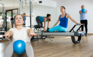 Concentrated elderly woman doing sit-ups with bender ball to strengthen abs muscles during group pilates class in fitness studio