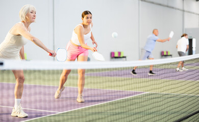 Two athletic women of different ages are playing a game of pickleball on a court inside a sports...