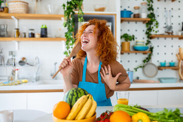 Funny beautiful woman singing into spatula, cooking in modern kitchen, holding spatula as microphone, dancing, listening to music, playful girl having fun with kitchenware, preparing food.