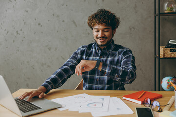 Successful smiling happy cheerful fun young employee business Indian man he wears casual blue checkered shirt looking at smart watch check time sit work at office desk with laptop pc computer indoors.