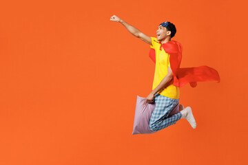 Full body side view young man wear pyjamas jam sleep eye mask coat rest relax home fly on pillow do super hero gesture isolated on plain orange background studio portrait Good mood night nap concept.