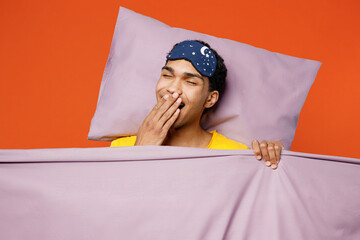 Calm young man wearing pyjamas jam sleep eye mask rest relax at home lies wrap covered under blanket duvet yawn isolated on plain orange color background studio portrait. Good mood night nap concept.