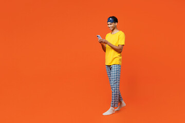 Fototapeta premium Full body side view young man wears pyjamas jam sleep eye mask rest relax at home hold in hand use mobile cell phone isolated on plain orange background studio portrait. Good mood night nap concept.