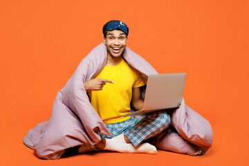 Full body young IT man in pyjamas jam sleep eye mask sit wrapped in duvet rest relax at home work...