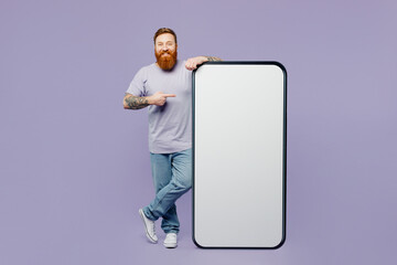 Full body young redhead man wear violet t-shirt casual clothes point index finger on big huge blank screen mobile cell phone smartphone with copy space mockup area isolated on plain purple background.