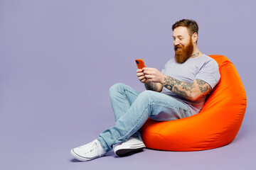 Full body young redhead bearded man wear violet t-shirt casual clothes sit in bag chair hold in...