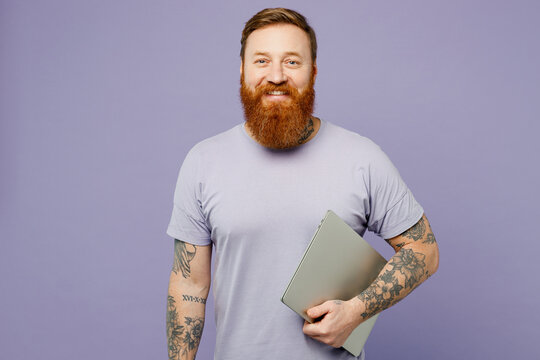 Young smiling happy redhead bearded IT man wearing violet t-shirt casual clothes holding closed laptop pc computer look aside isolated on plain pastel light purple background studio Lifestyle concept.