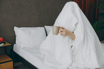 Young man sitting on bed covered with duvet wrapped in blanket drink coffee tea hiding be afraid of...