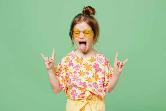 Little child kid girl 6-7 years old wear casual clothes sunglasses do horns up gesture, depicting heavy metal rock sign isolated on plain green background. Mother's Day love family lifestyle concept.
