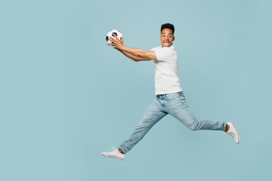 Full body side profile view fun young man fan wears t-shirt cheer up support football sport team jump high hold catch soccer ball watch tv live stream isolated on plain pastel blue color background.