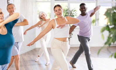 Fototapeta premium Young girl coach oversees mentoring group of elderly clients in fitness studio. Multiracial people group of different ages practice repeating dance movements