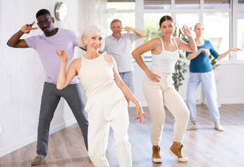 Fototapeta premium ..Young woman teacher teaches group of multiethnic mature active people dancing modern dance moves in studio