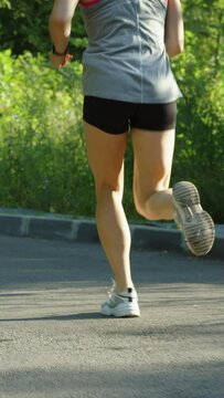 Vertical Screen: Female feet wearing sports sneakers running on asphalt road on sunny morning in slow motion. Tracking shot partial view of woman training outside. Concept of healthy lifestyle