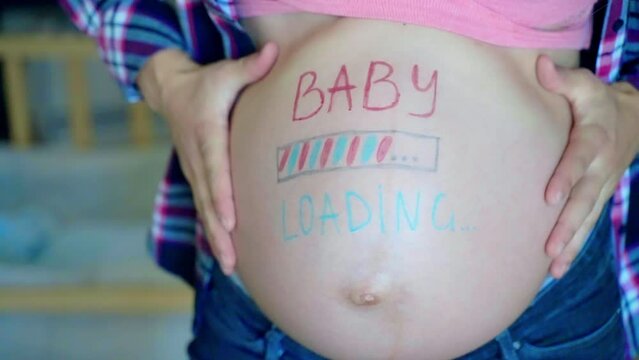 Blue and pink baby loading inscription on woman's pregnant belly, expecting mom waiting for her baby arrival, happy female slowly posing on camera and showing her thumbs up