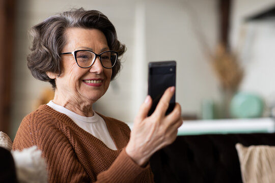 Happy Mature Middle Aged Woman Enjoying Using Mobile Apps, Texting, Sitting On Sofa, Smiling Senior Woman Holding Smart Phone, Looking At Mobile Phone Screen, Browsing Social Media At Home