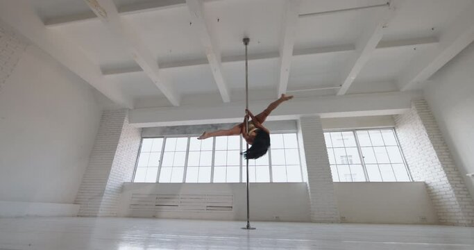 Asian strip dancer spinning on a pylon, in white daylight studio against the background of panoramic window in beige lingerie. Slow motion. Seductive flexible gorgeous perfect sensual performance
