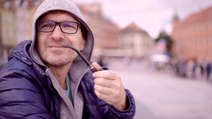Portrait of adult man with glasses sitting in the hood on square and smoking a tobacco pipe in the Palace Square, Warsaw Old Town