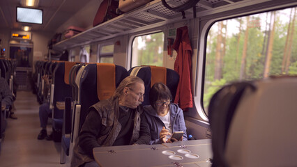Elderly couple travel by train, the lady is holding a mobile phone in her hand, both are looking at the smartphone and talking with each other
