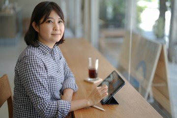 Short hair Asian woman is sitting at table to using digital tablet and smart pen to search...