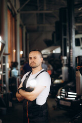 Fototapeta premium Handsome Car Mechanic is Posing in a Car Service. He Wears a Jeans Shirt and Safety Glasses. His Arms are Crossed. Specialist Looks at a Camera and Smiles