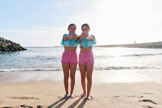 Twin sisters in matching outfits holding squirt guns on beach at sunny day