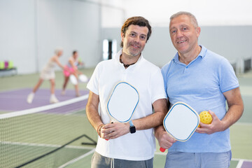 Two positive smiling men in sportswear with rackets and balls in hands posing near net on indoor pickleball court after friendly match..