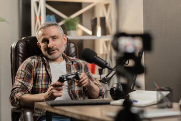 A gray-haired man in a plaid shirt sits in front of the camera and holds a gamepad in his hands. A male tech blogger is recording a video review of a new gamepad.