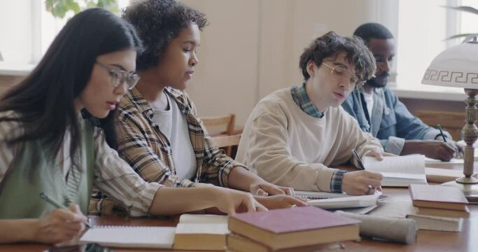 Multiethnic group of students men and women studying together in university classroom writing reading book and talking. Teamwork and educational project concept.