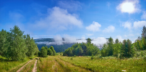 Fototapeta na wymiar Panorama of the mountainous countryside near the forest. Morning fog melts under the rays of the bright summer sun. Dirt road invites for a morning walk
